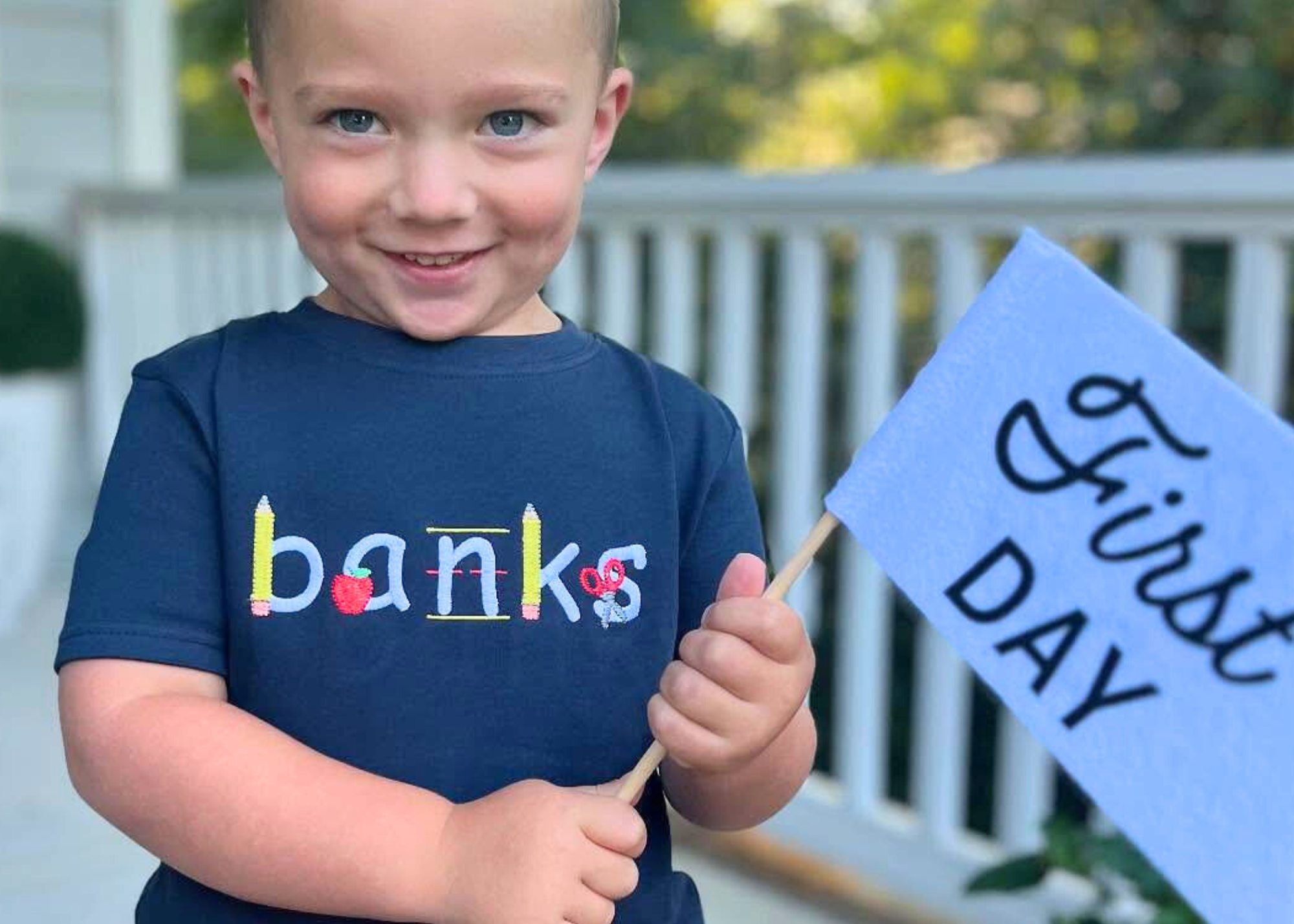 A child wearing a navy blue t-shirt with the name 'banks' embroidered in colorful letters, holding a sign that reads '1st DAY'.