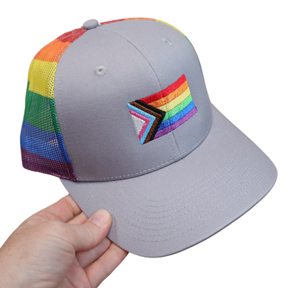 Gray structured snapback hat with the Progress Pride flag machine embroidered on the front and a rainbow striped mesh back, held in hand